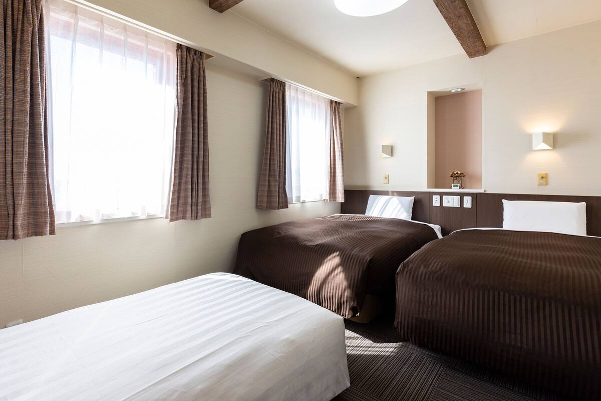 Habitación de hotel con dos camas, un ventanal con cortinas, una lámpara en el techo, una mesita de noche con lámpara y un espejo en la pared.