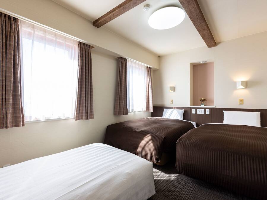 Ein Hotelzimmer mit zwei Betten, einem großen Fenster mit Vorhängen, einer Deckenleuchte, einem Nachttisch mit Lampe und einem Spiegel an der Wand.