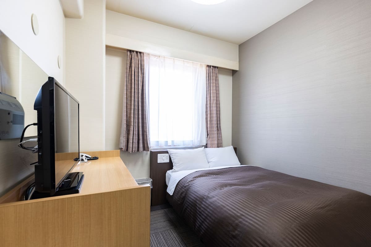 Habitación de hotel con cama, escritorio con computadora, ventana con cortinas y una lámpara fijada a la pared.