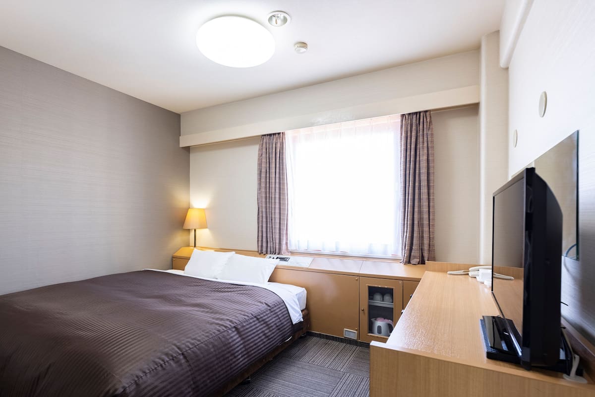 Habitación de hotel con cama, escritorio, televisor, lámpara y ventana con cortinas.