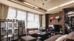 Una sala moderna con un sofá, un reposapiés y un televisor. Se observa una estantería con diversos objetos y una ventana con cortinas.