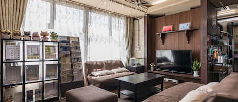 Una sala moderna con un sofá, un reposapiés y un televisor. Se observa una estantería con diversos objetos y una ventana con cortinas.