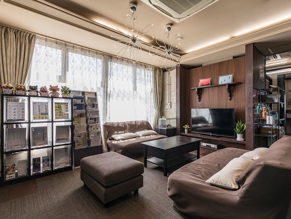 Una sala moderna con un sofá, un reposapiés y un televisor. Se observa una estantería con diversos objetos y una ventana con cortinas.