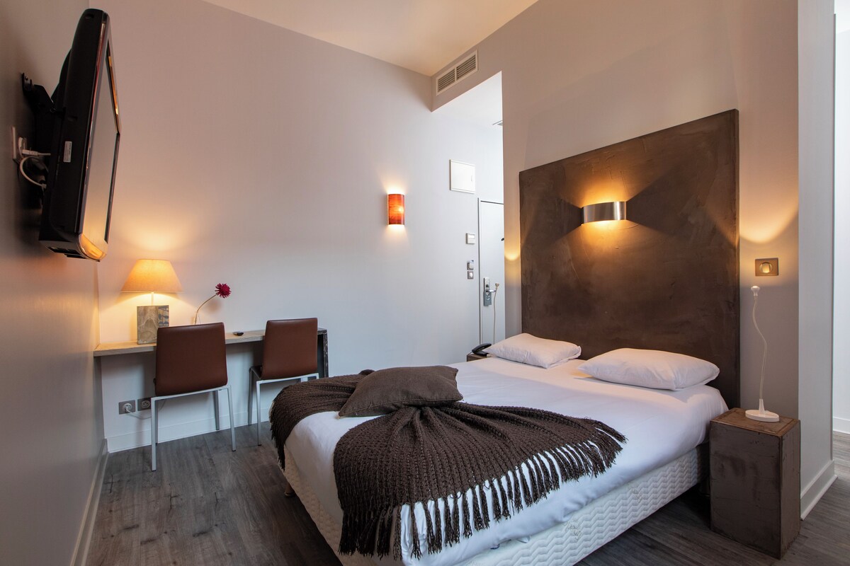 A modern hotel room with a large bed, a desk with two chairs, a wall-mounted TV, and a bedside table with a lamp.