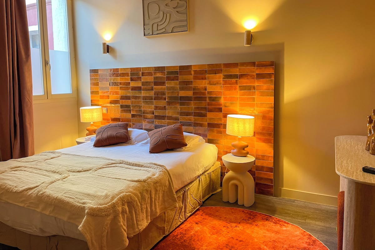 A hotel room with a bed, two bedside lamps, a headboard with a mosaic design, a small round table with a lamp, and a bathroom area with a sink and mirror.