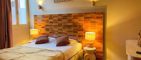 A hotel room with a bed, two bedside lamps, a headboard with a mosaic design, a small round table with a lamp, and a bathroom area with a sink and mirror.