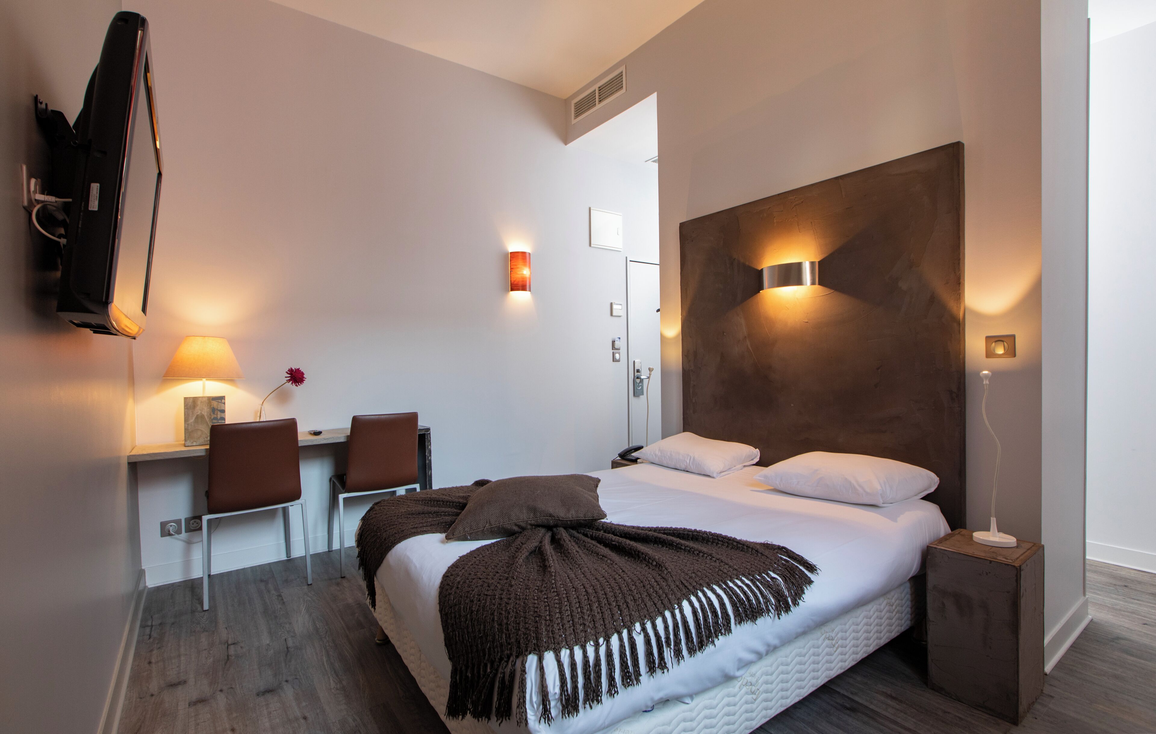Una habitación de hotel moderna con una cama grande, un escritorio con dos sillas, un televisor montado en la pared y una mesita de noche.