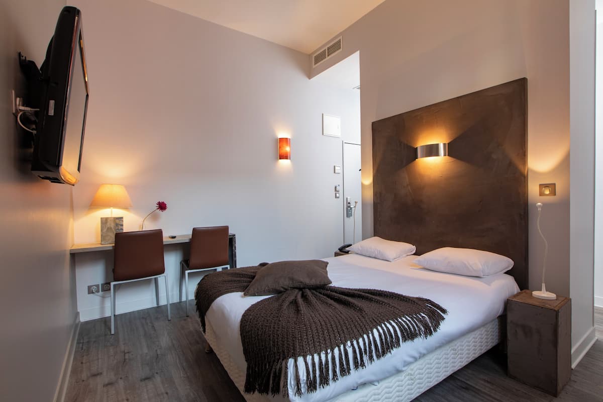 A modern hotel room with a large bed, a desk with two chairs, a wall-mounted TV, and a bedside table with a lamp.