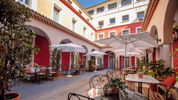 An outdoor café area with tables and chairs, surrounded by potted plants and umbrellas.