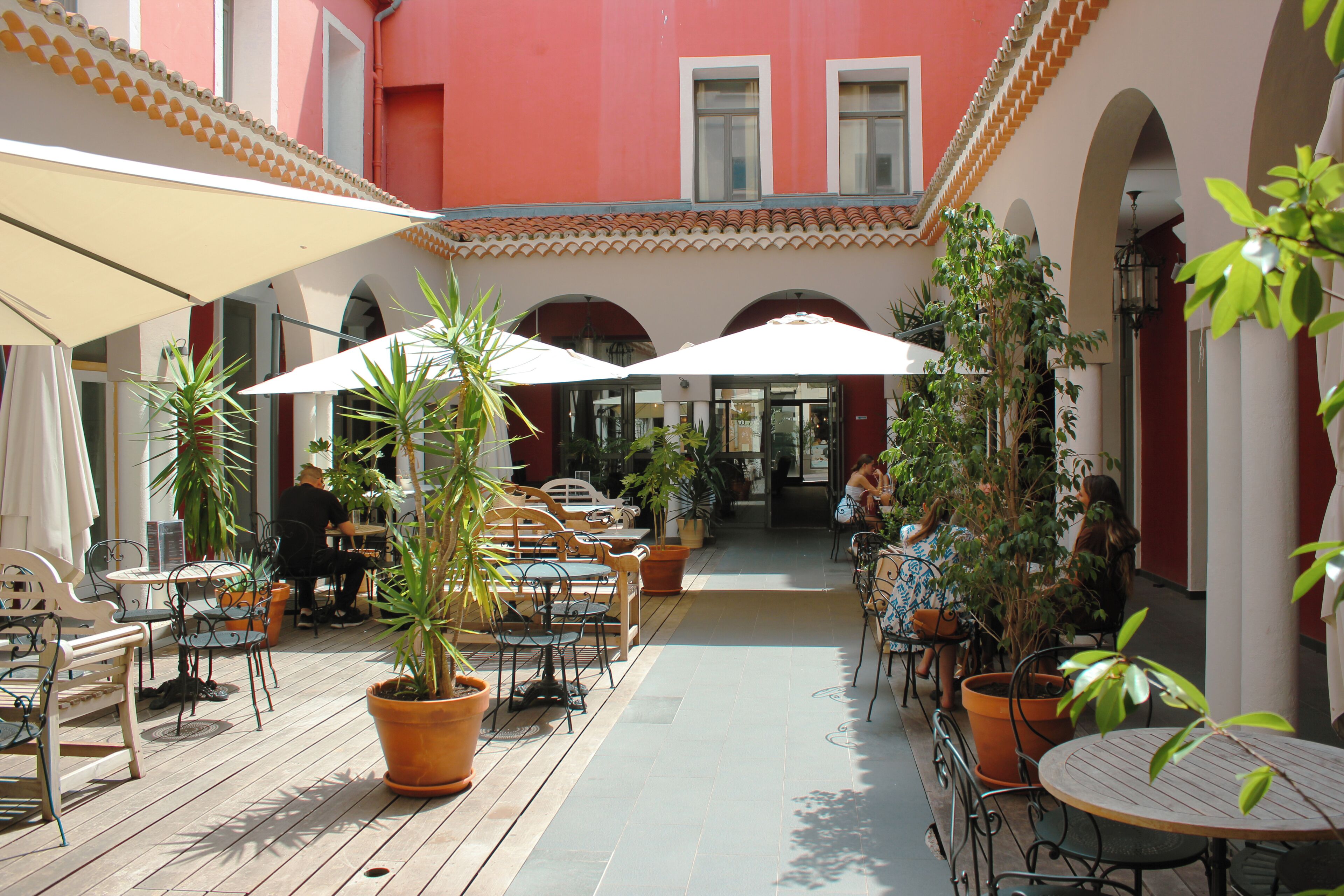 Un café en plein air avec des tables et des chaises, des plantes en pot et un sol carrelé.