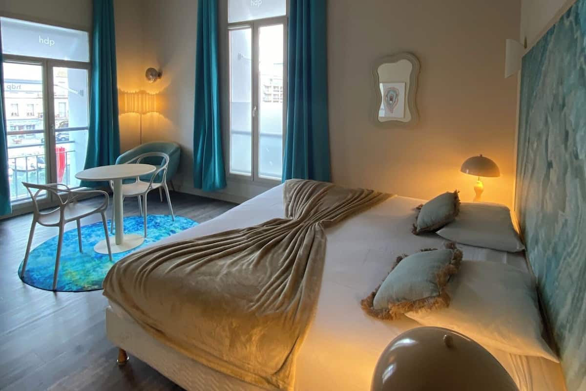 A hotel room with a bed, a desk, and a chair. There is a window with curtains, a lamp, and a painting on the wall.
