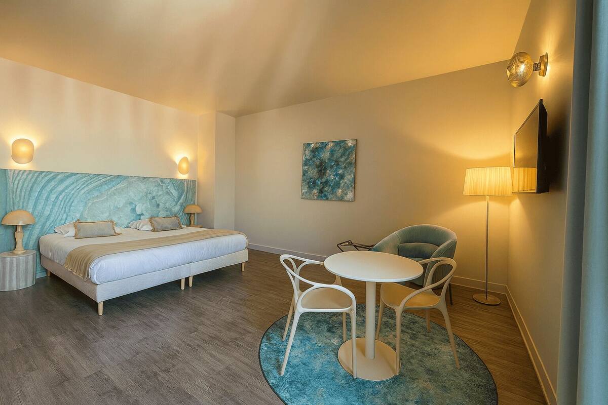 A modern hotel room with a bed, bedside tables, a desk with chairs, a painting, and a TV on the wall.