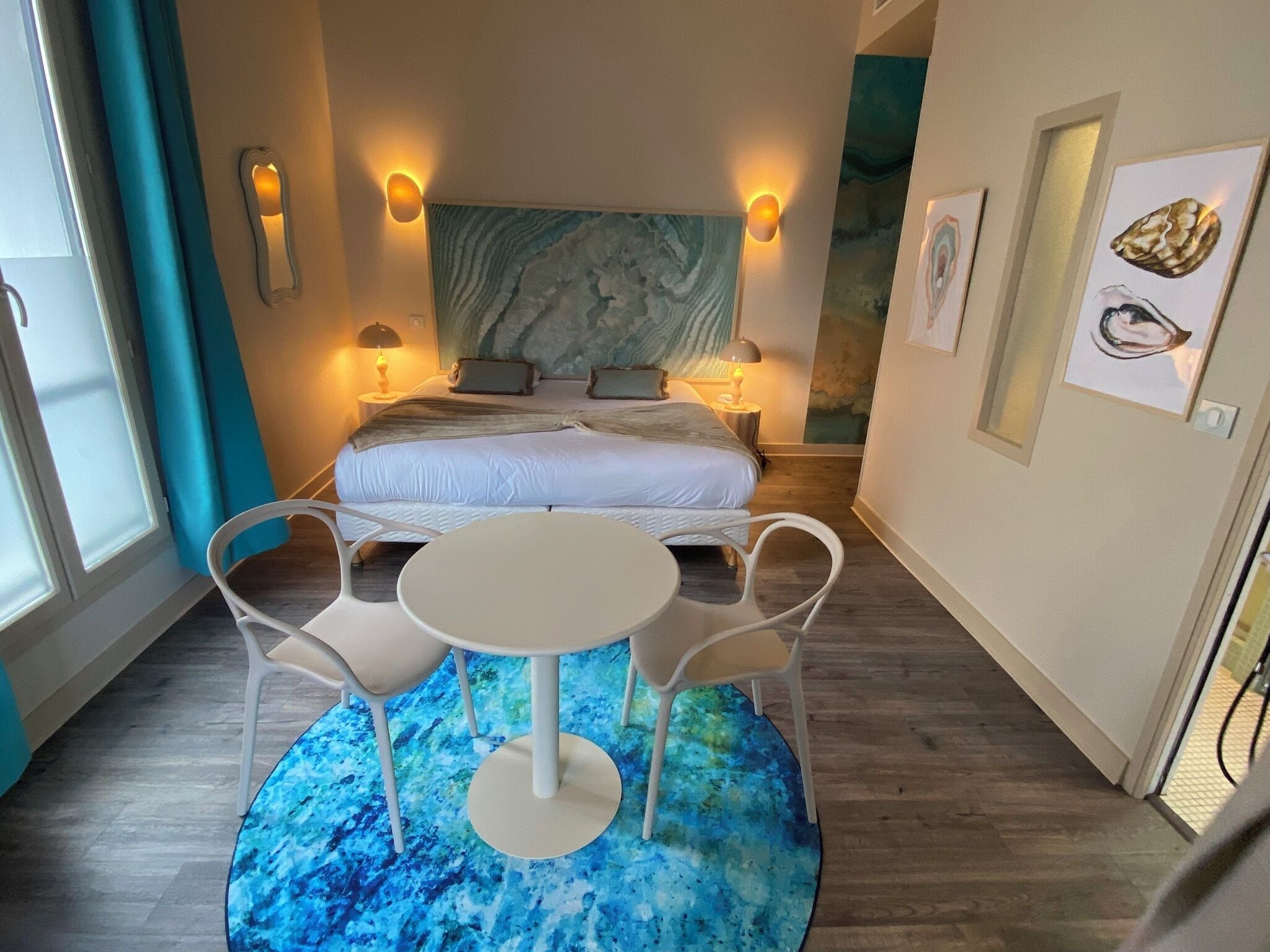 A bedroom with a bed, two bedside tables with lamps, a round table with two chairs, and a blue patterned rug.