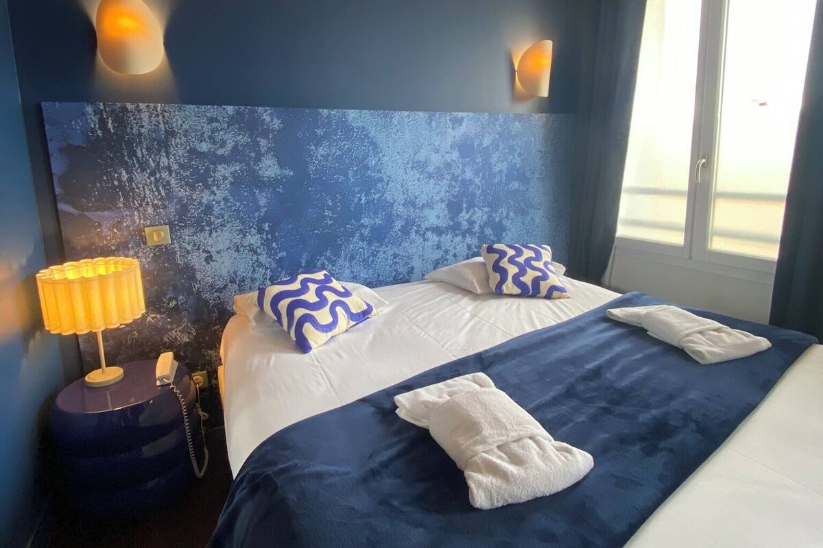 A hotel room with a large bed, two wall-mounted lamps, a bedside telephone, and a window with curtains.