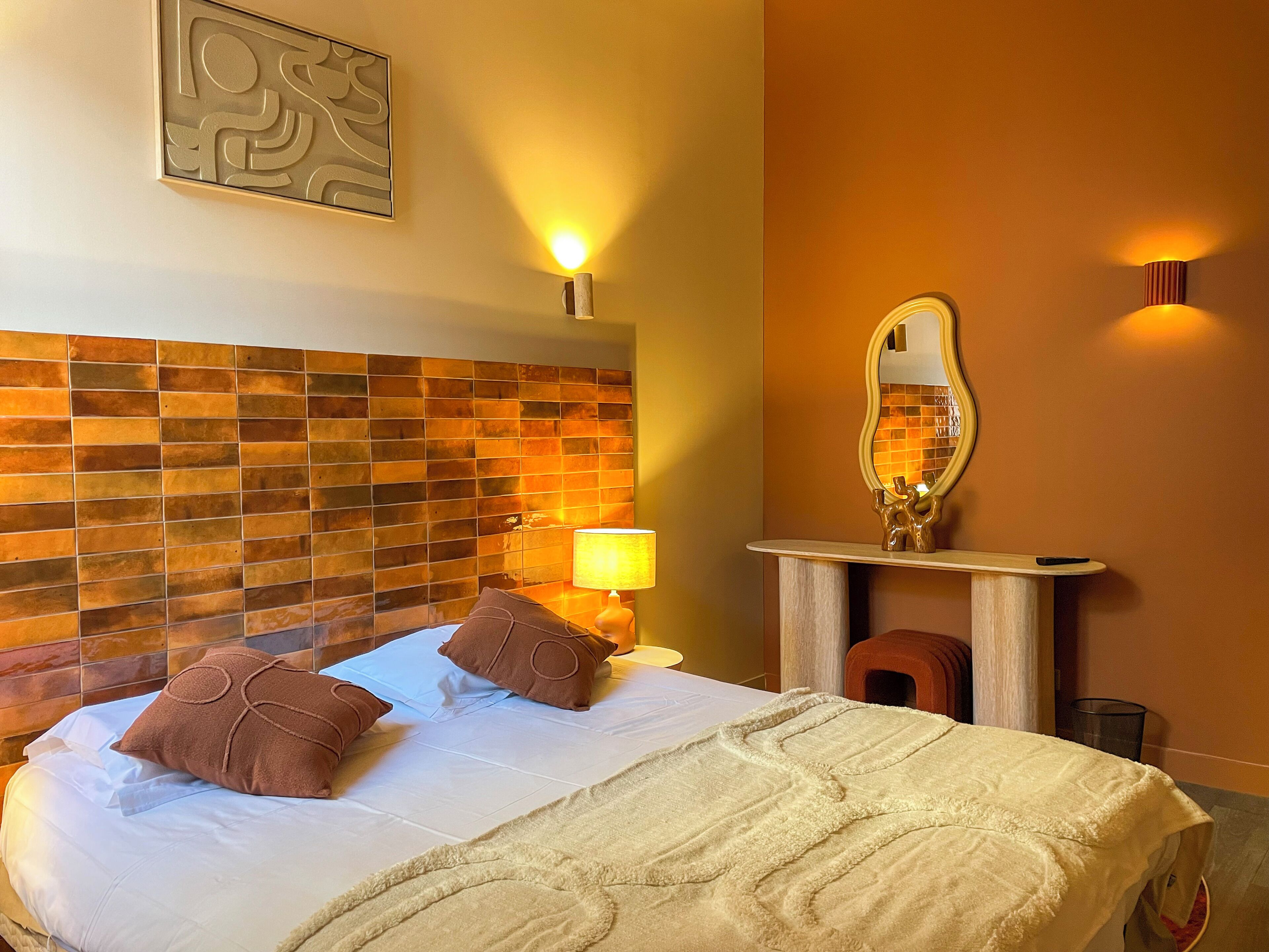 A hotel room with a tiled headboard, a bed with white linens and brown pillows, a bedside table with a lamp, and a mirror above the table.