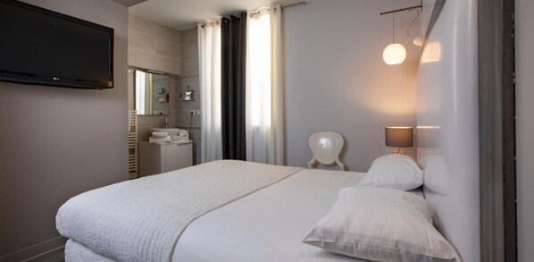 A hotel room with a bed, a TV mounted on the wall, a bathroom with a sink and mirror, and a chair. - Hotel de Paris (Sète)