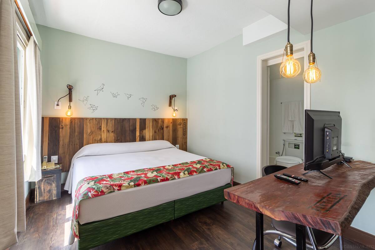 Un dormitorio con cabecera de madera, cama con cubrecama floral, escritorio con computadora y control remoto, y lámpara de pared.