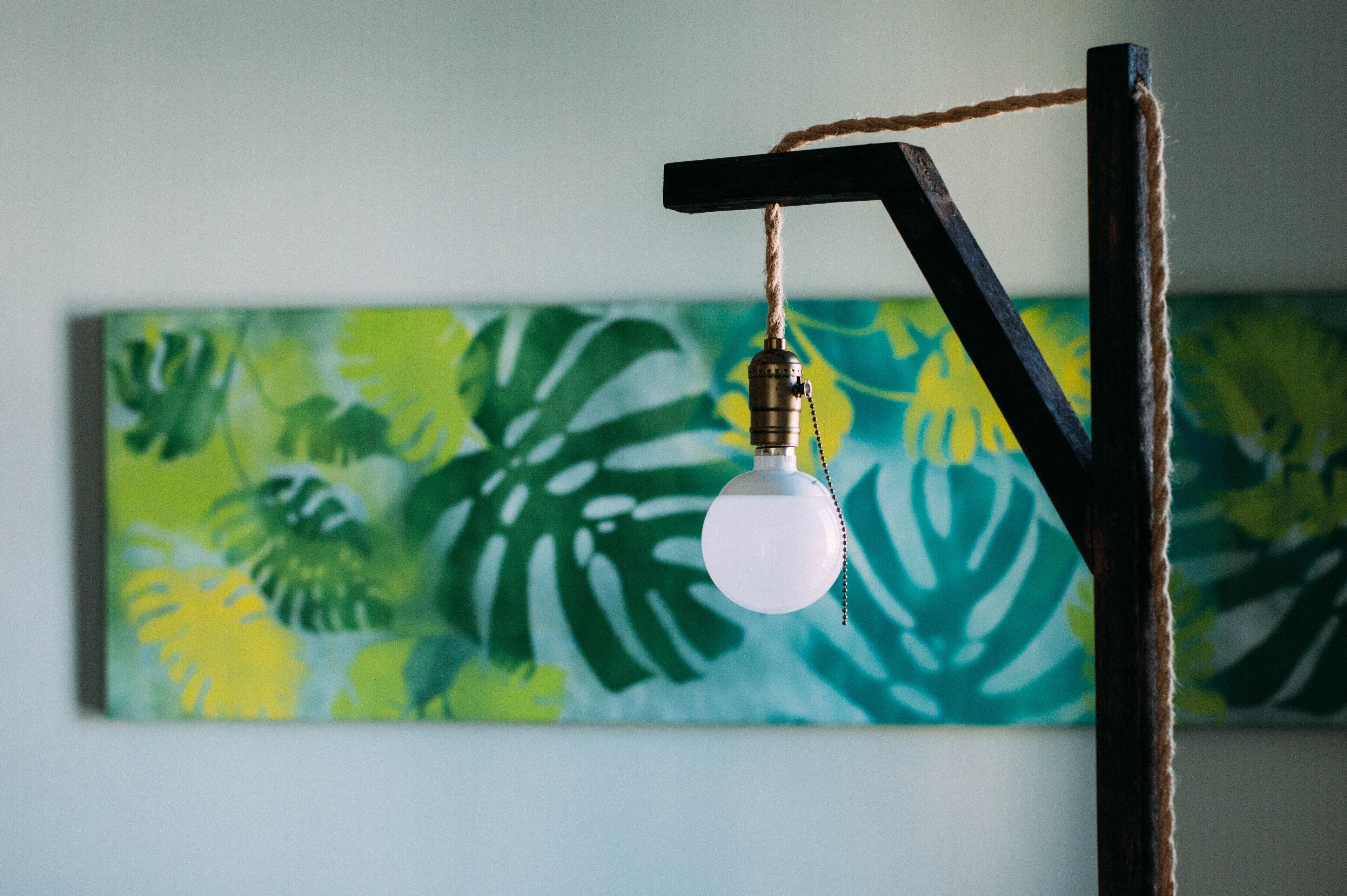 A light bulb hanging from a wooden structure against a wall with a tropical leaf painting.