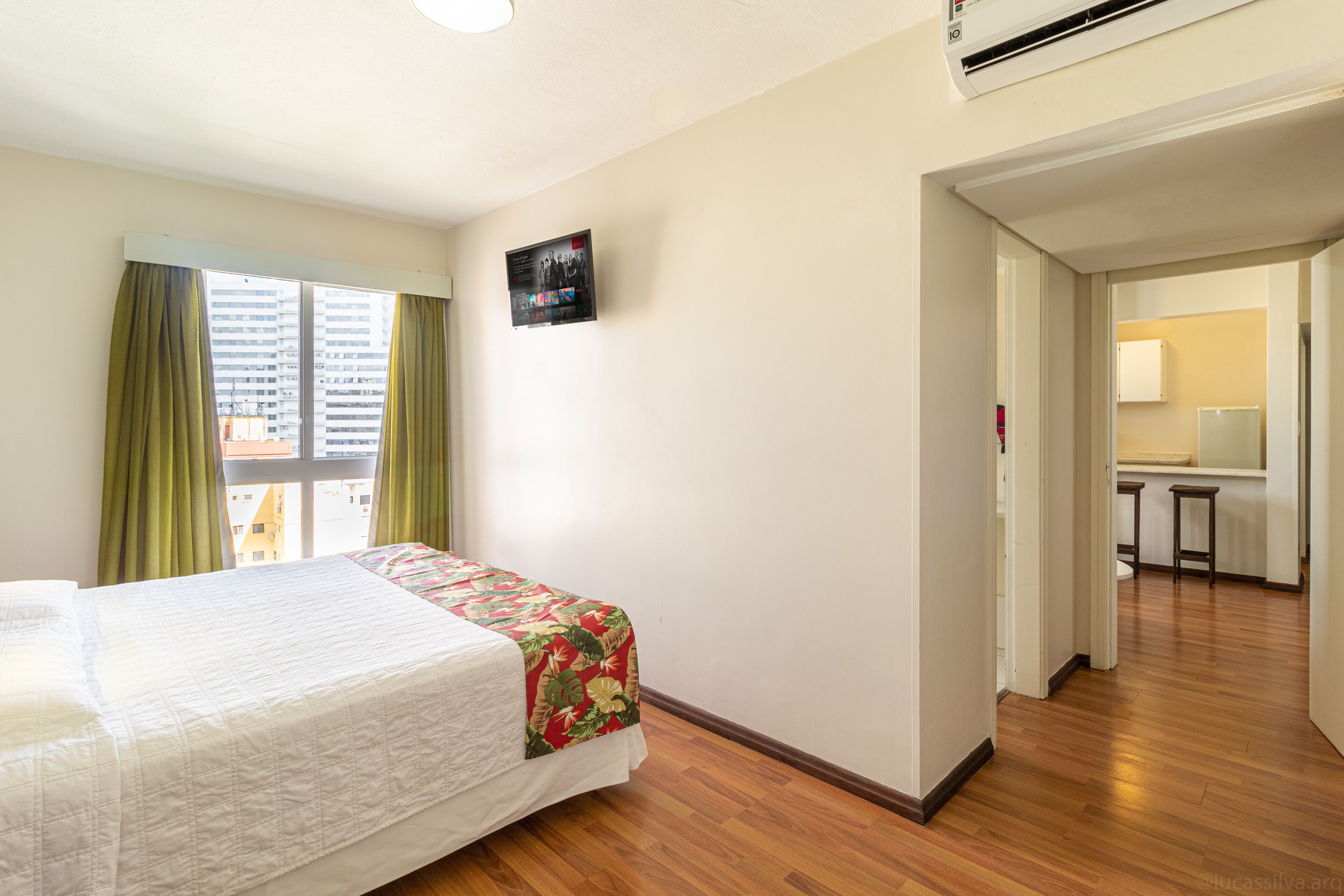 Quarto de hotel com cama, janela com cortinas, pequena cozinha e banheiro visível por uma porta aberta.