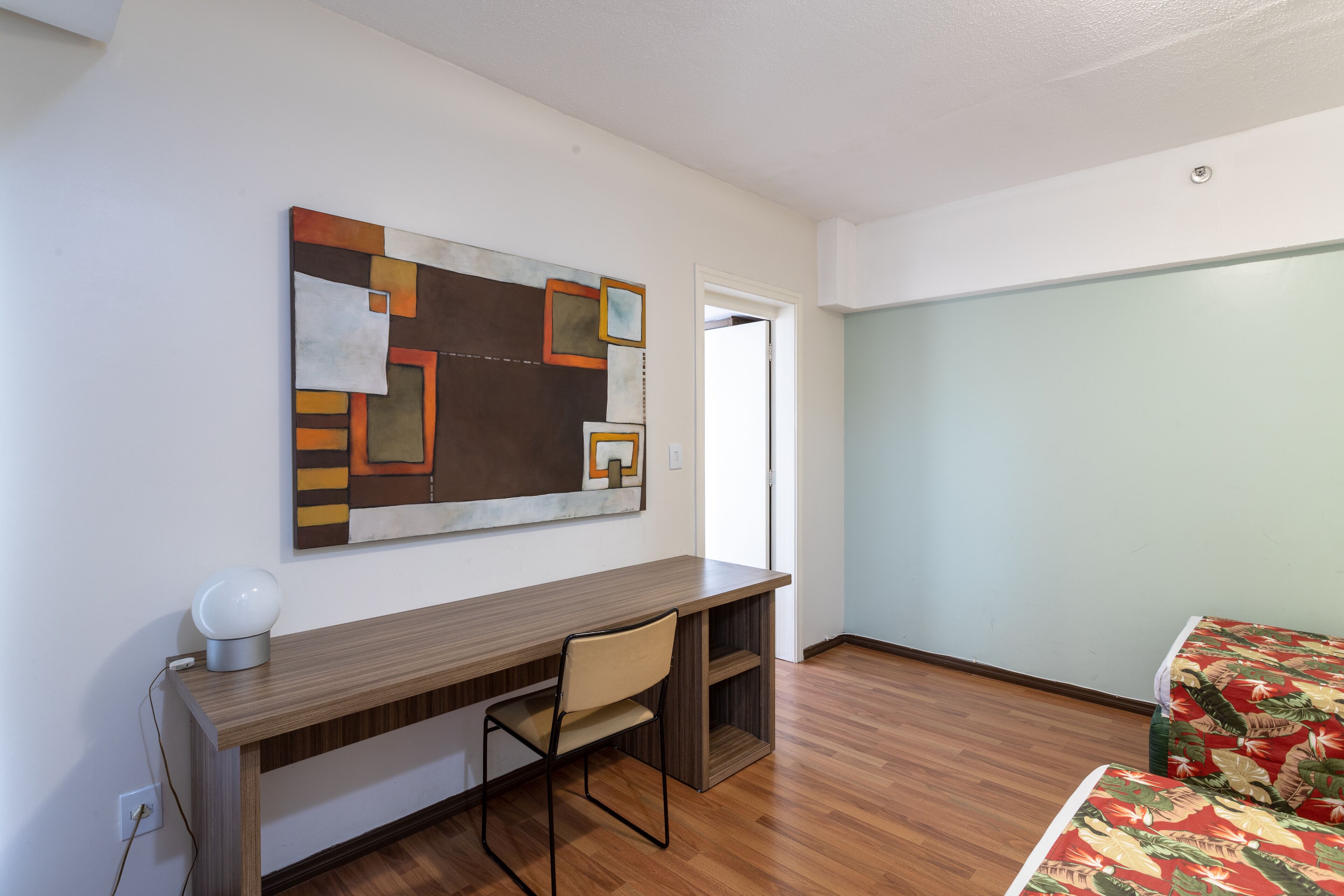 Quarto com escrivaninha de madeira, uma cadeira, cama com colcha florida e parede com arte abstrata.