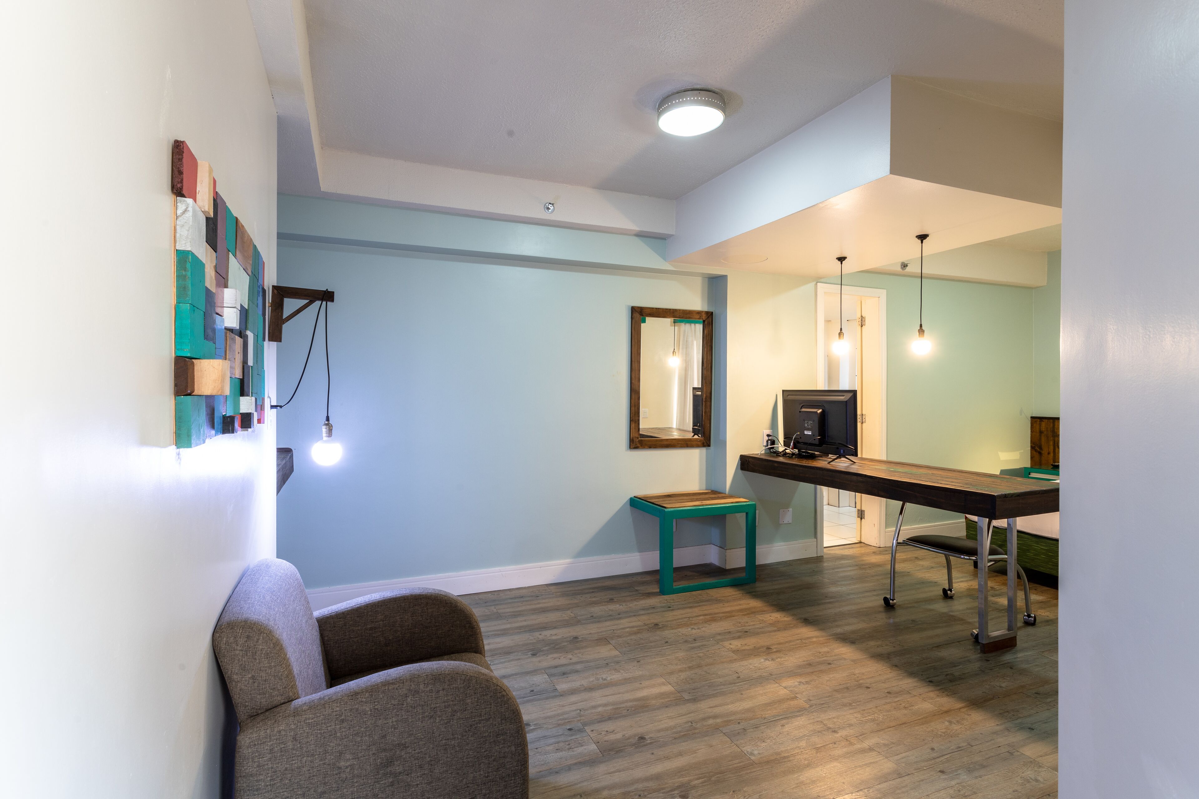 Una habitación moderna con piso de madera, un escritorio con computadora, una silla, un taburete pequeño y un aplique de pared.