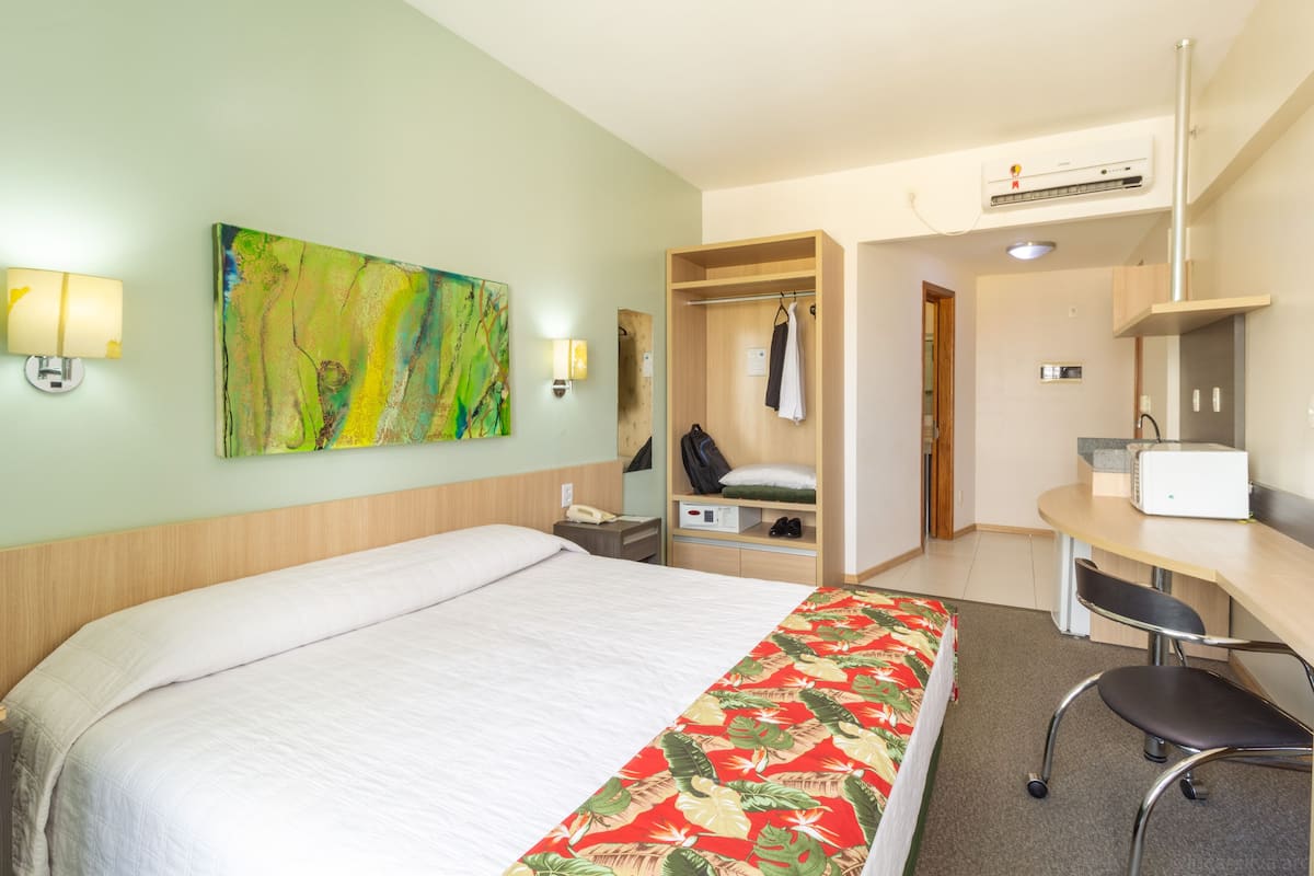 Habitación de hotel con cama, escritorio, silla, armario y un cuadro en la pared.