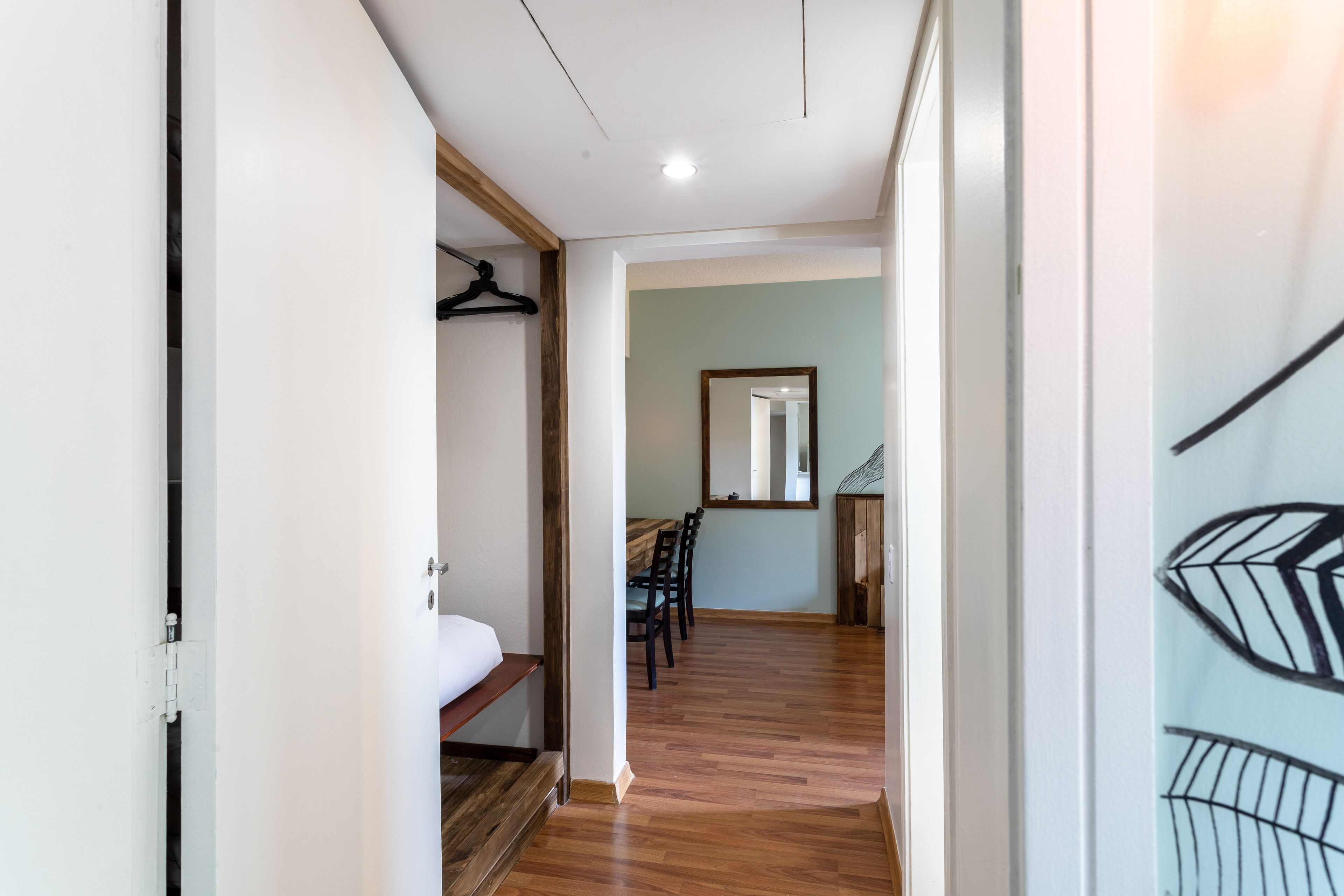 A hallway with a bed, a wooden bench, a dining table with chairs, and a mirror on the wall.