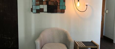 A room with a light-colored armchair, a small wooden side table, and a wall-mounted light fixture.