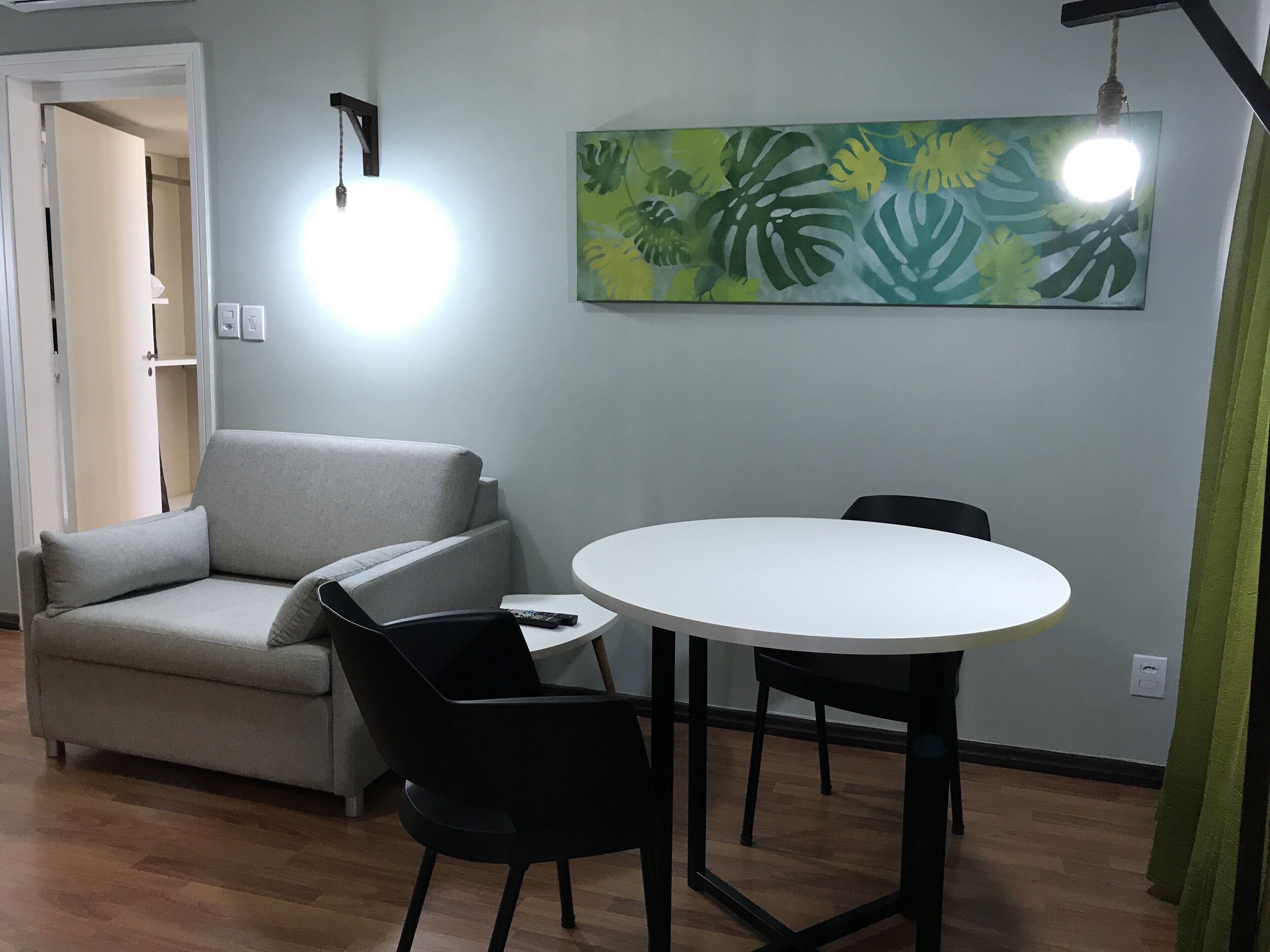 Un salón con un sofá gris, una mesa redonda blanca y dos sillas negras. Hay un cuadro con hojas tropicales en la pared y una lámpara colgante.