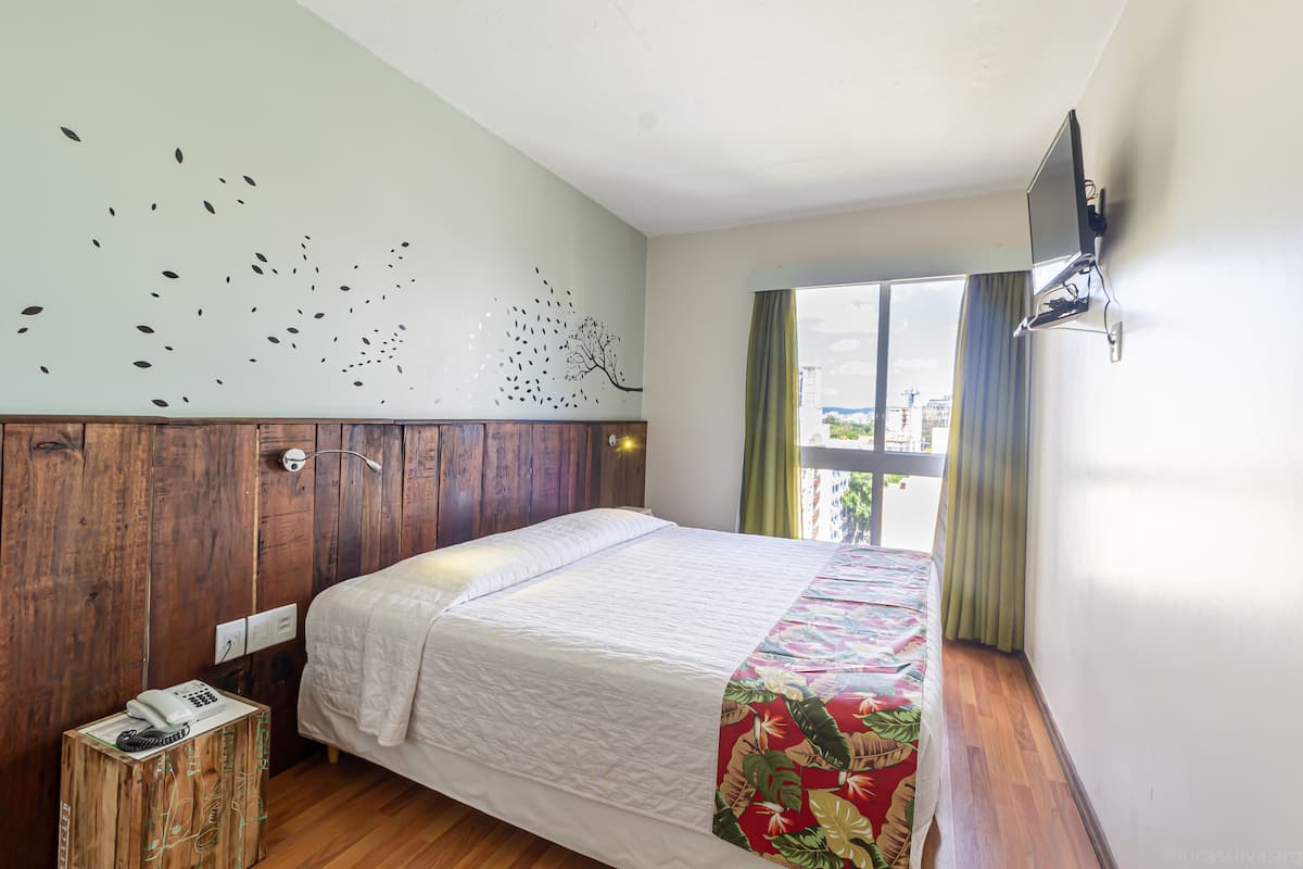 Habitación de hotel con cama, cabecera de madera, televisor montado en la pared, ventana con cortinas y una pequeña mesa de madera con un teléfono.