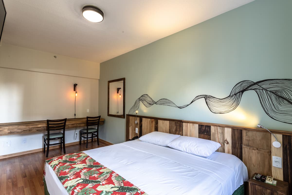 Habitación de hotel con cabecera de madera, cama con ropa blanca, colcha de estampado floral, escritorio con dos sillas y una lámpara de pared.