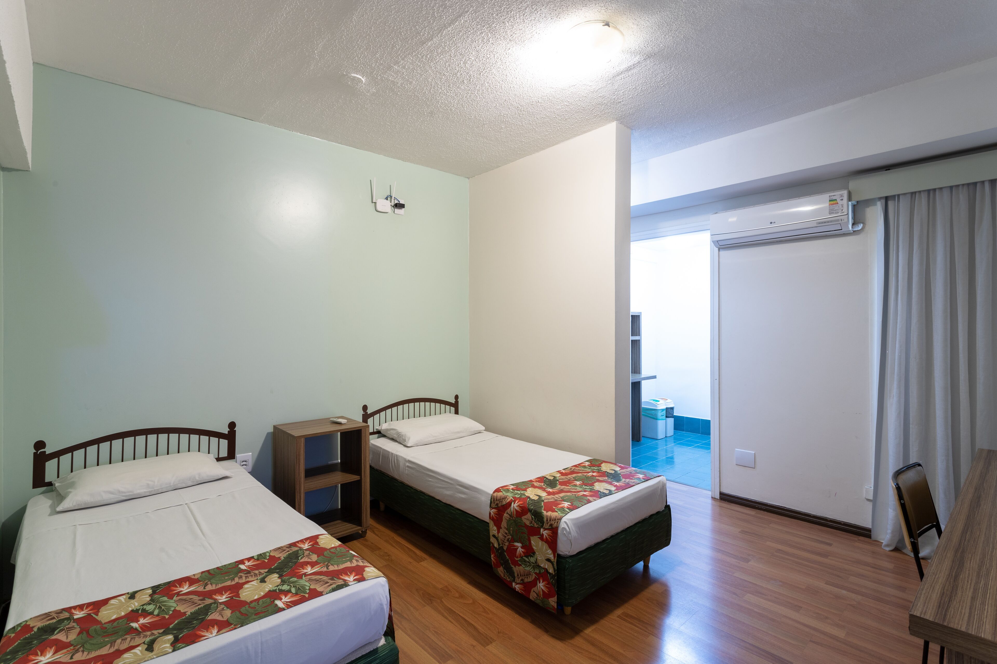 A hotel room with two single beds, a wooden desk, a chair, a small shelf, and a sliding door leading to a balcony.