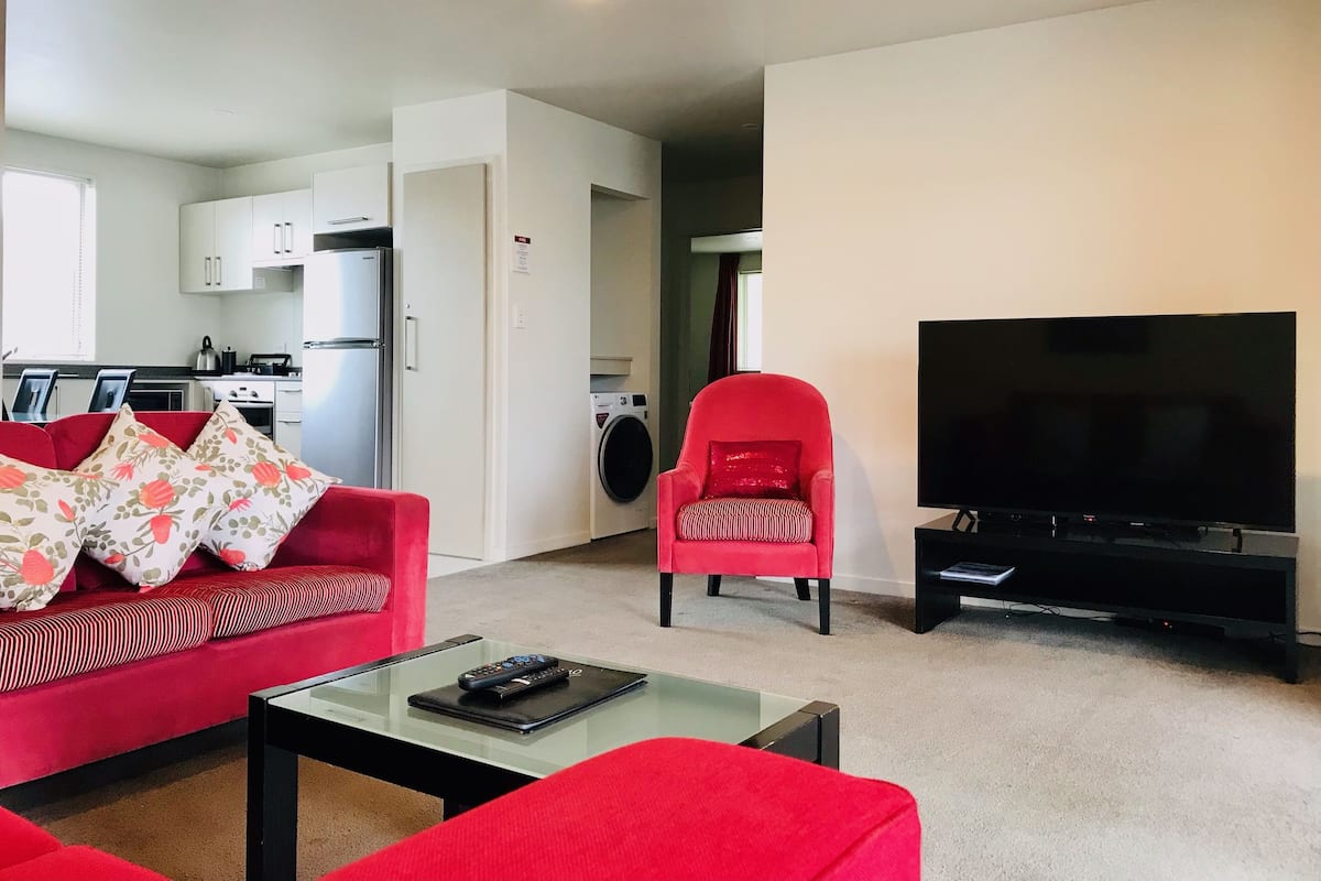 Een woonkamer met een rode bank, een rode fauteuil, een glazen salontafel, een flatscreen televisie en een wasmachine op de achtergrond.