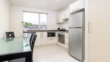 Cozinha moderna com armários brancos, geladeira de aço inoxidável, micro-ondas e forno. Há uma janela com uma planta no parapeito, e uma mesa de jantar com cadeiras.