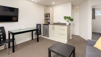 A compact living space with a kitchenette, a dining area with a glass table, a black sofa, and a flat-screen TV mounted on the wall.