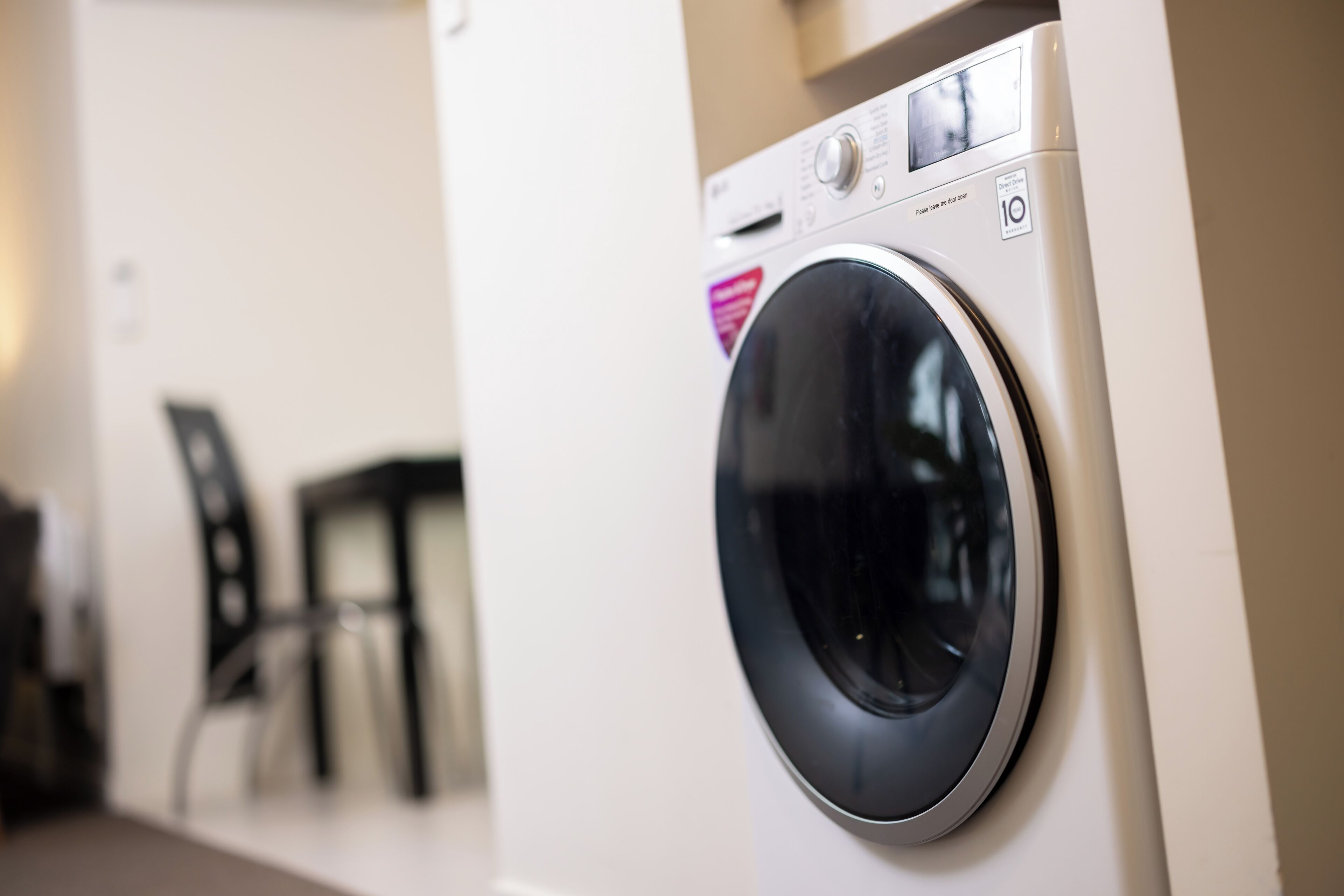 A front-loading washing machine in a room with a white wall and a chair in the background.