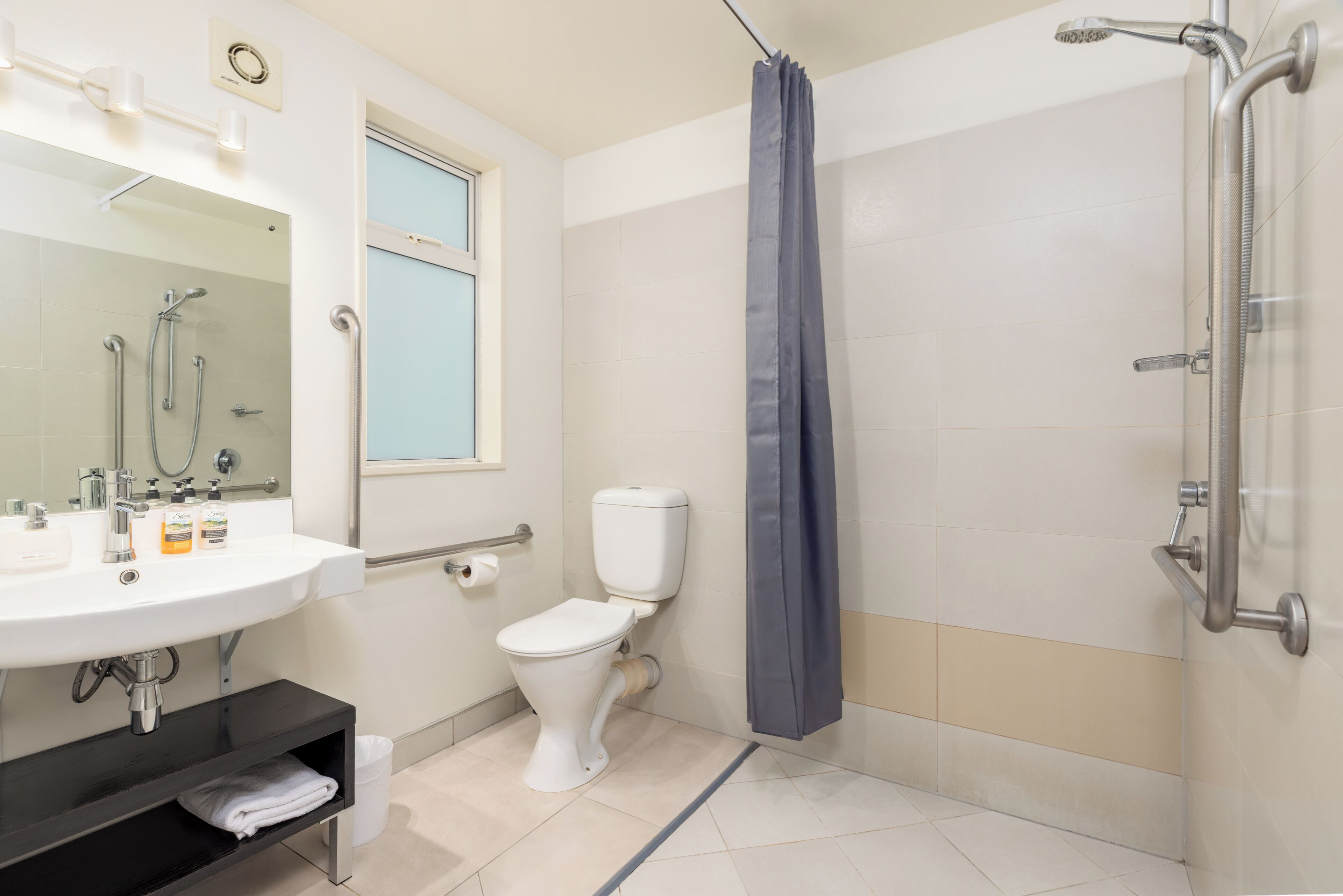 Ein modernes Badezimmer mit einer weißen Toilette, einem Waschbecken mit Seifenspendern, einer Dusche mit Duschbrause und einem Fenster mit Vorhang.