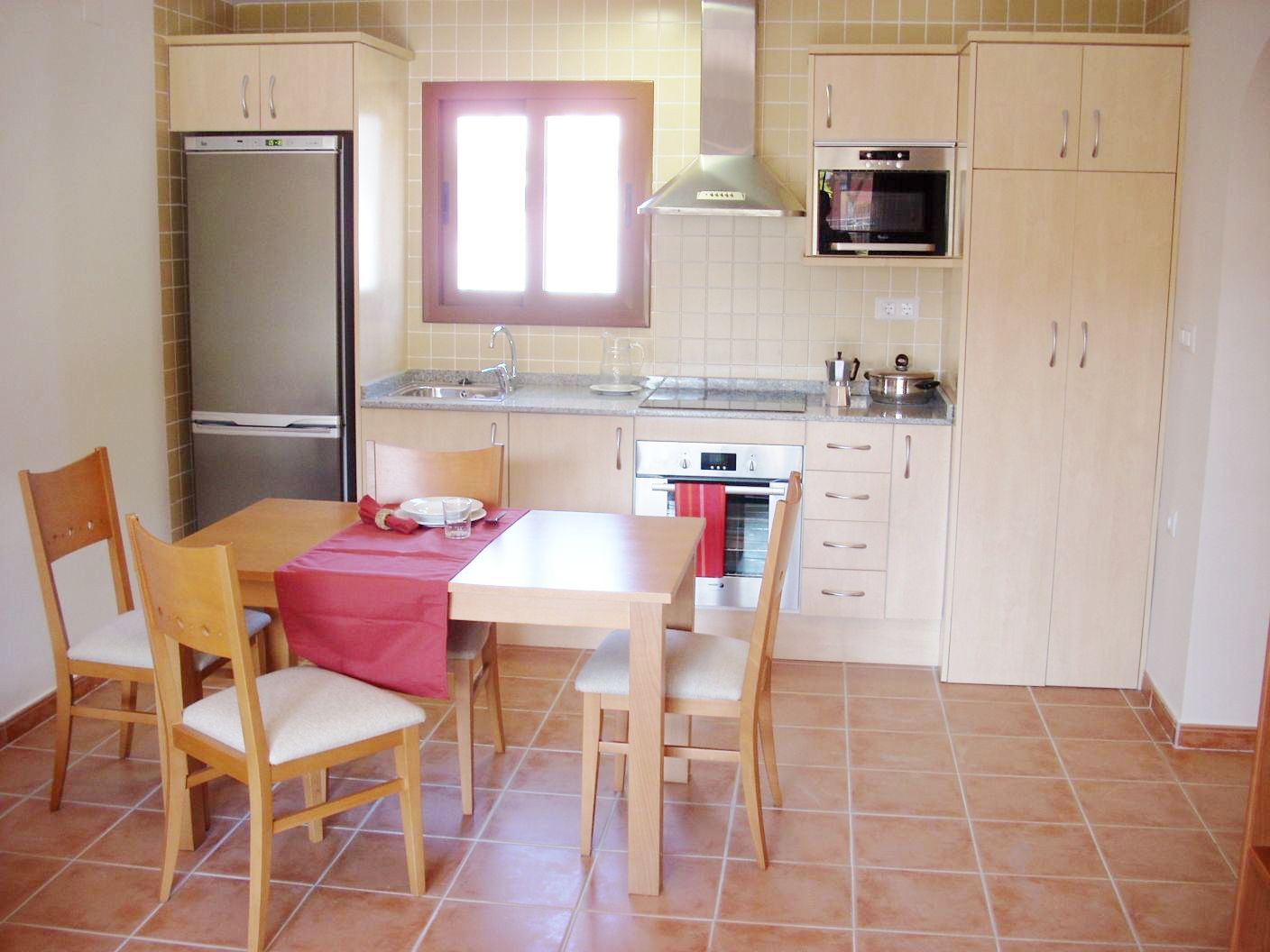A compact kitchen with a refrigerator, oven, and sink. There are two wooden chairs and a table with a red tablecloth.