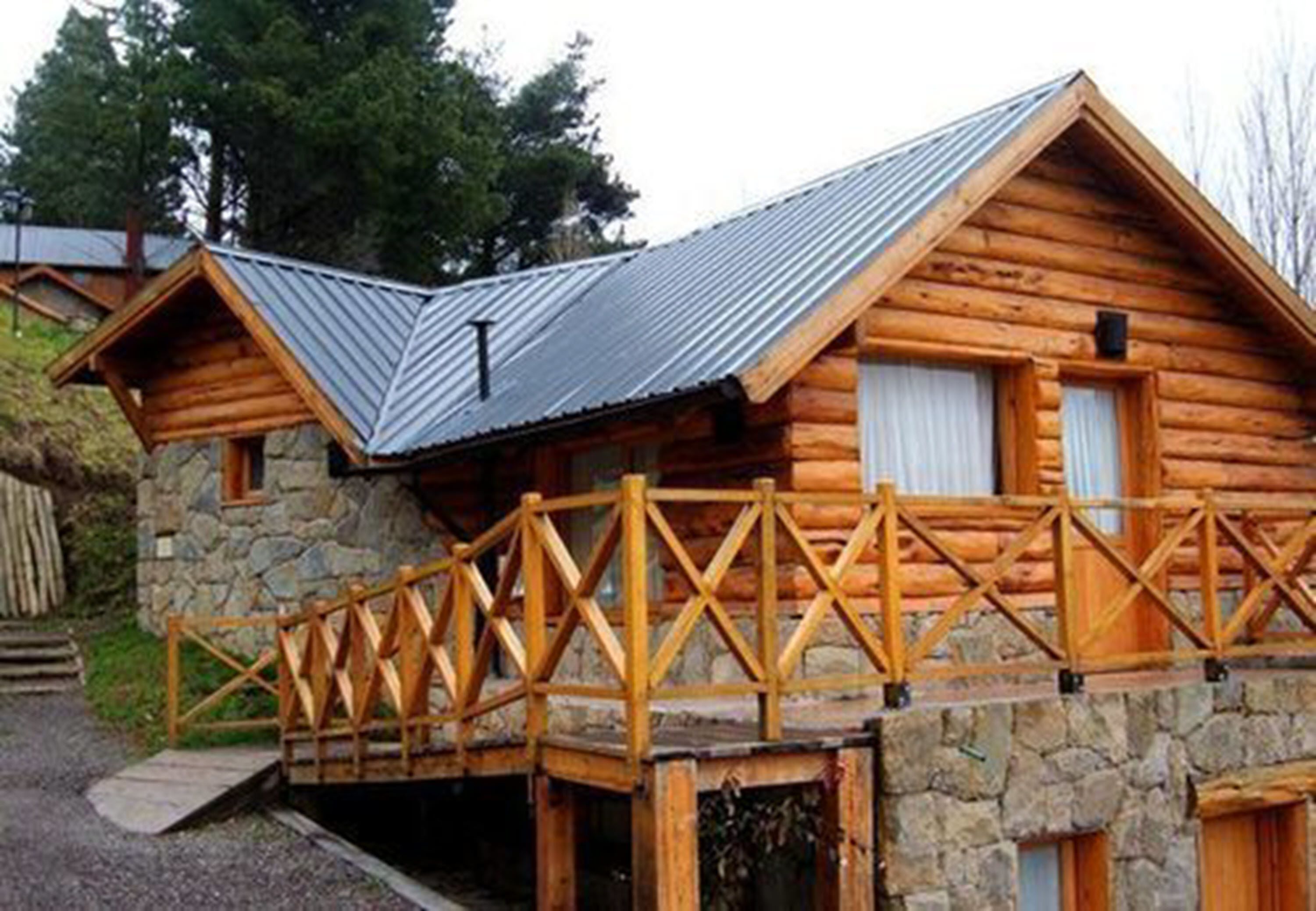 Una cabaña de troncos con cimientos de piedra, techo de metal y terraza de madera con barandilla.