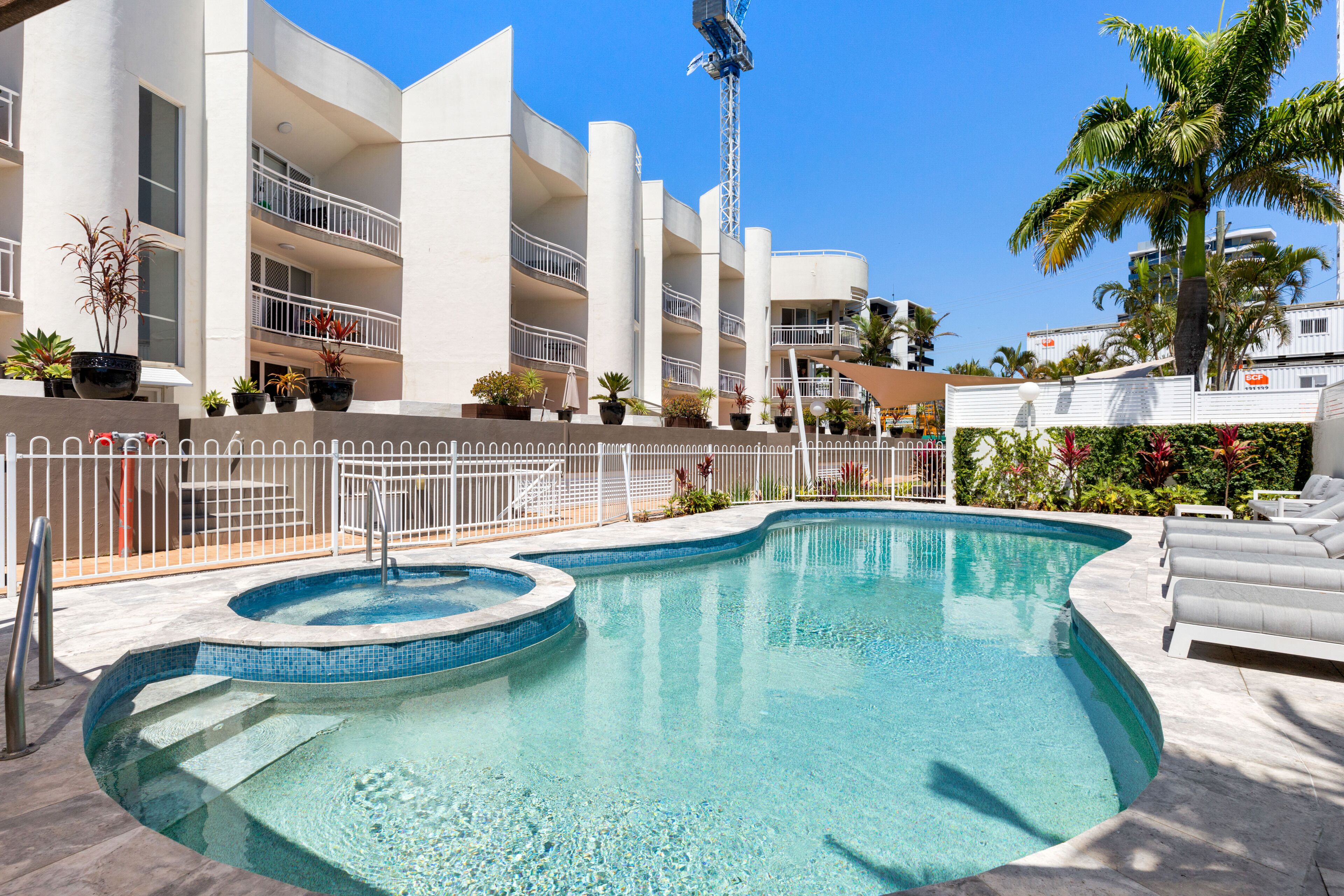 A swimming pool with a small hot tub, surrounded by a white fence, and a building with balconies and potted plants in the background.