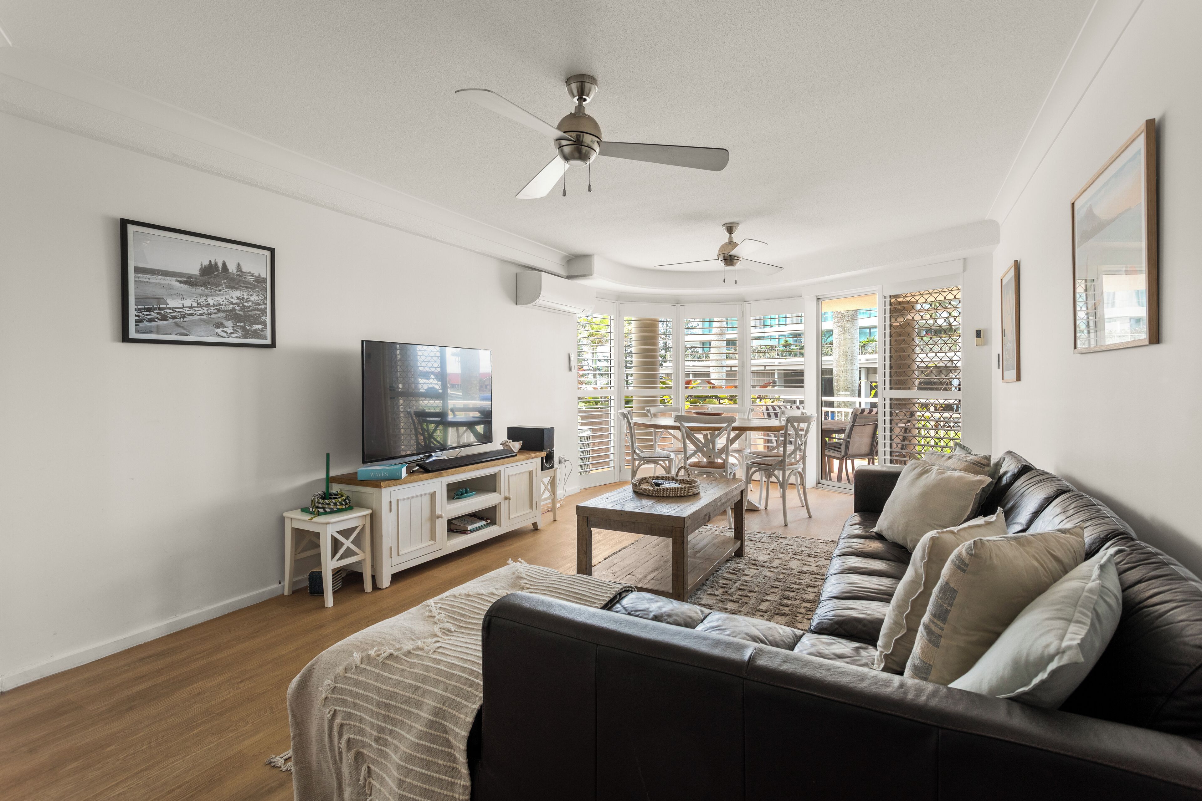 Un salon moderne avec un canapé en cuir, une table basse et un téléviseur posé sur un meuble blanc. On y trouve également un ventilateur de plafond, un tableau encadré au mur et une vue sur l’extérieur grâce à de grandes fenêtres.
