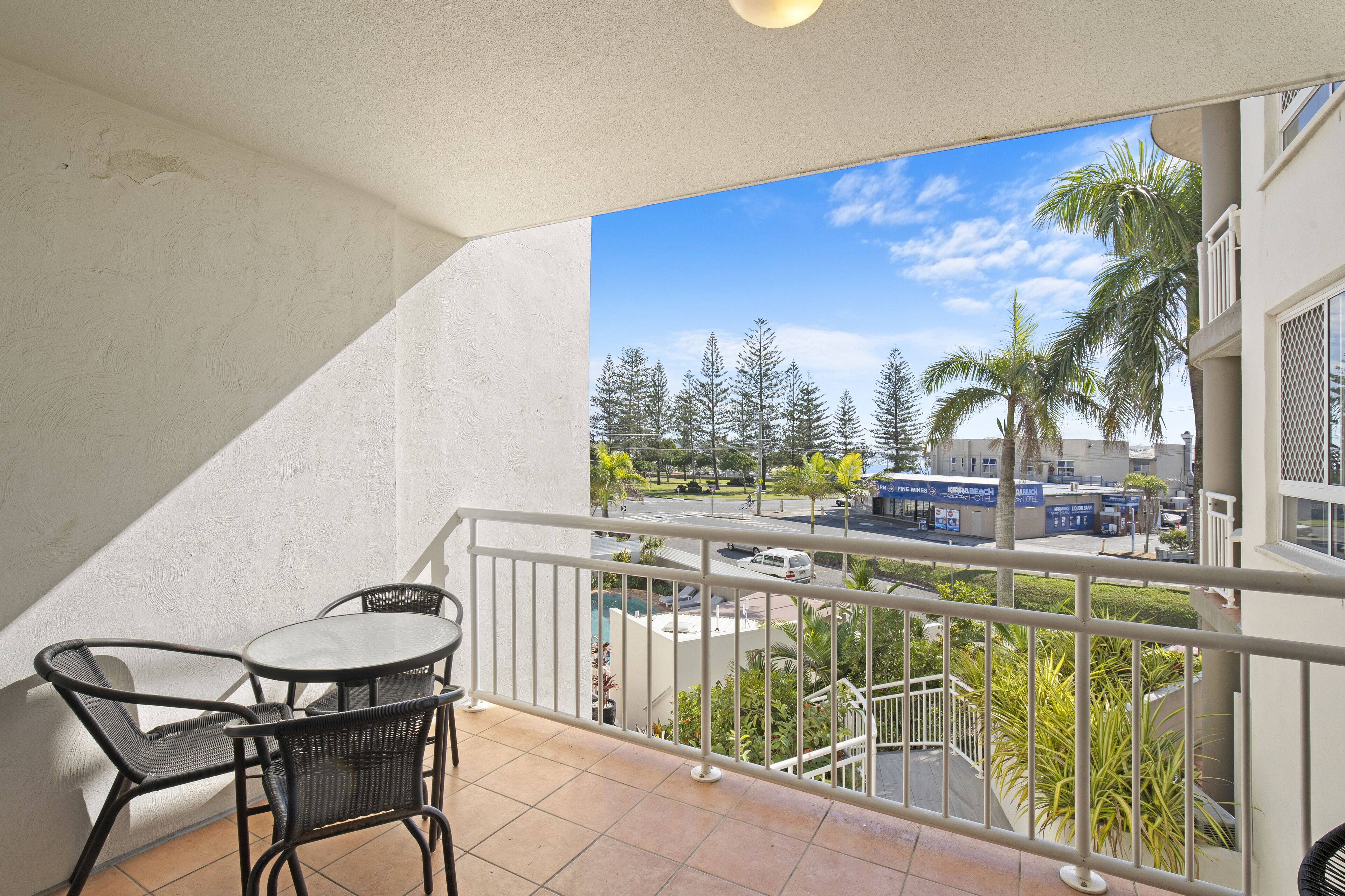 A balcony with a table and chairs, overlooking a street with palm trees and buildings.