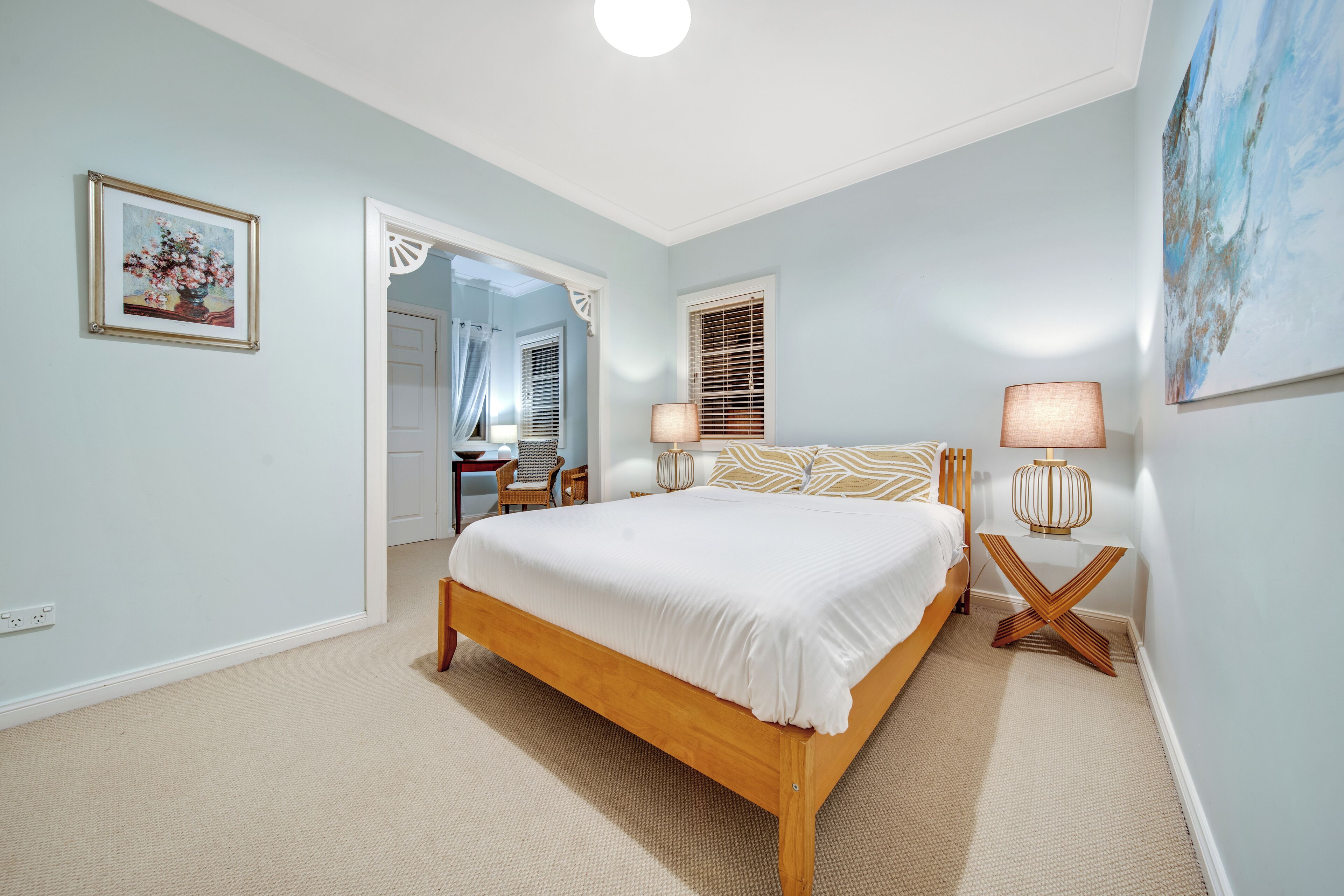 Quarto com cama de madeira, duas luminárias de cabeceira, um quadro e uma porta que dá para outro cômodo.