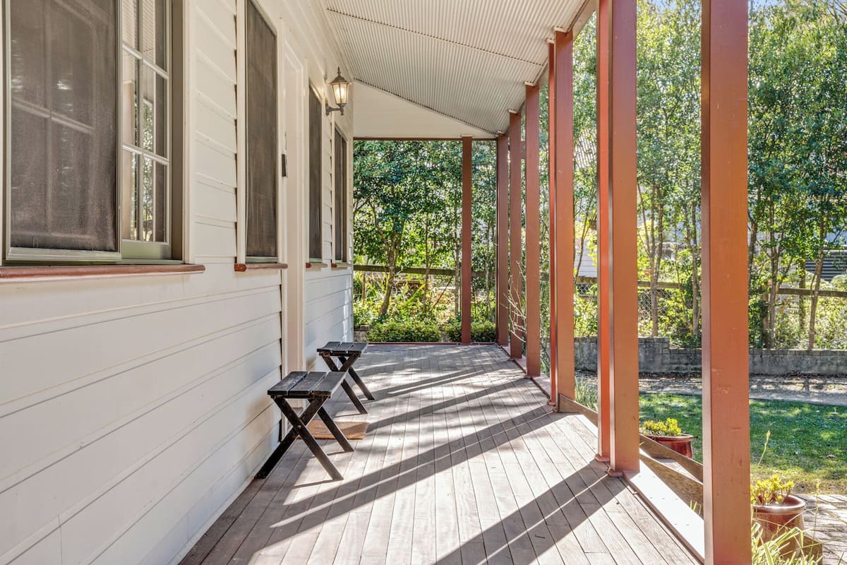 A covered porch with two wooden benches, a white exterior wall, and a view of a garden.