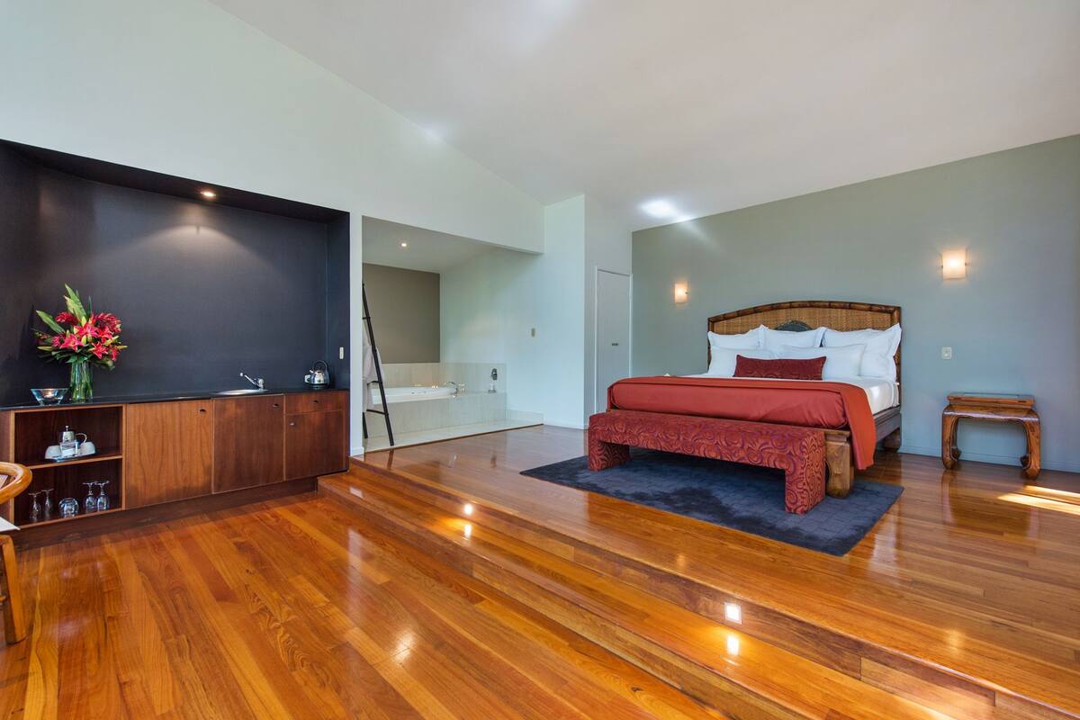 Un dormitorio moderno con suelo de madera, una cama grande con ropa de cama roja, una mesita de noche de madera y una pequeña mesa.