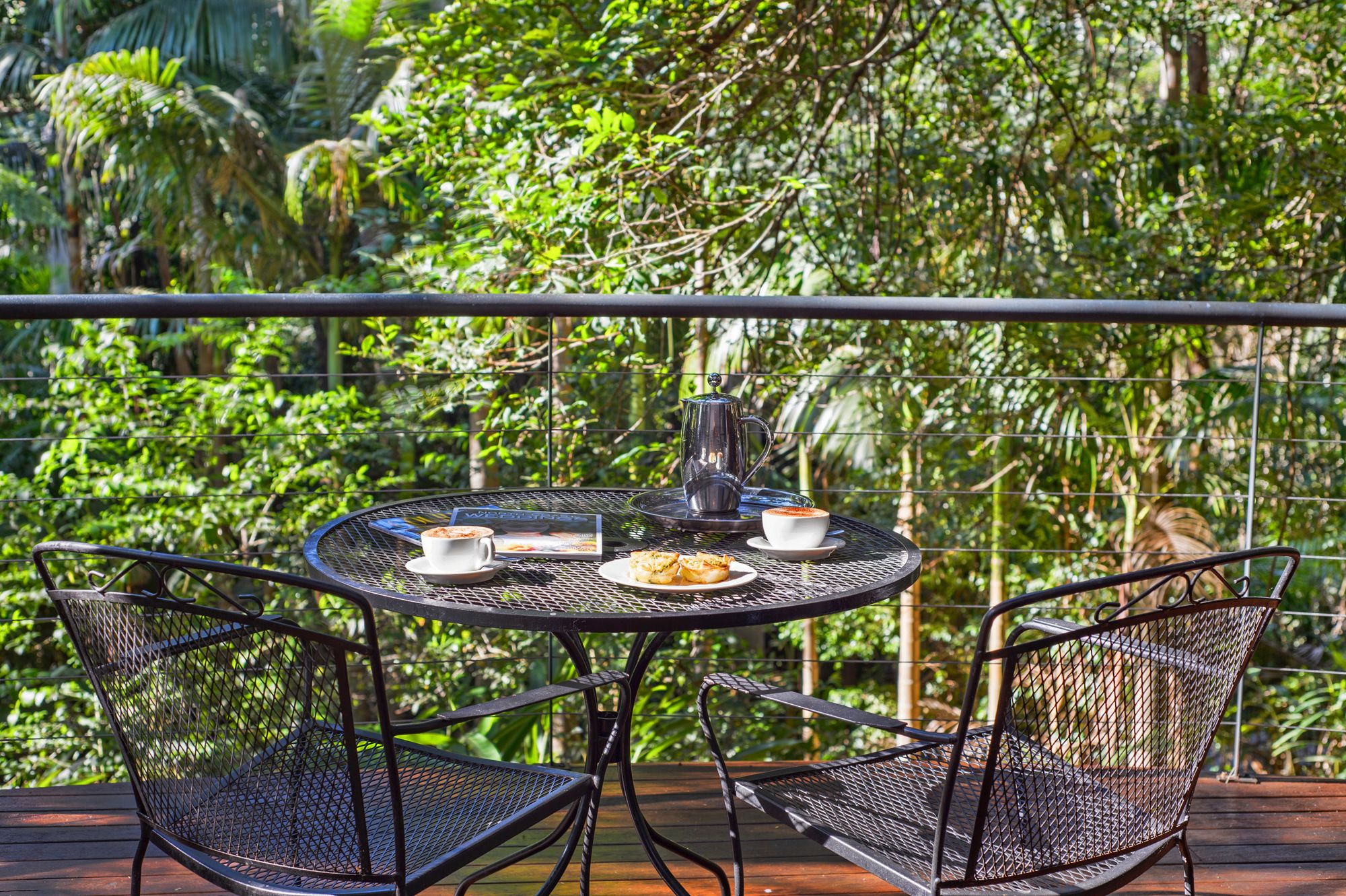 A round metal table with chairs, a coffee cup, a plate with pastries, and a coffee pot on a balcony with a metal railing.