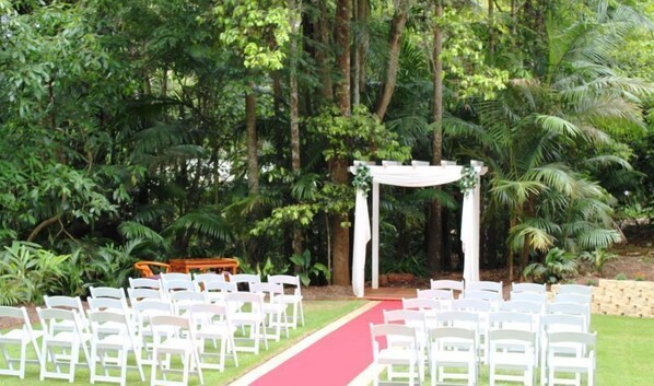 Un espacio al aire libre con jardín, sillas blancas dispuestas en filas, una alfombra roja y un arco blanco.
