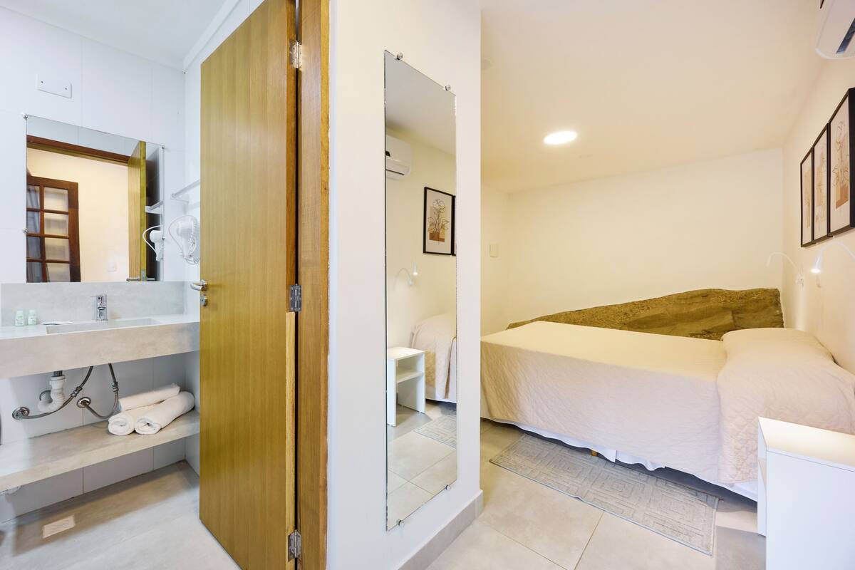A hotel room with a wooden door, a white bed, a bedside table, a white sink, and a mirror.