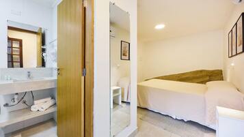 A hotel room with a wooden door, a white bed, a bedside table, a white sink, and a mirror.
