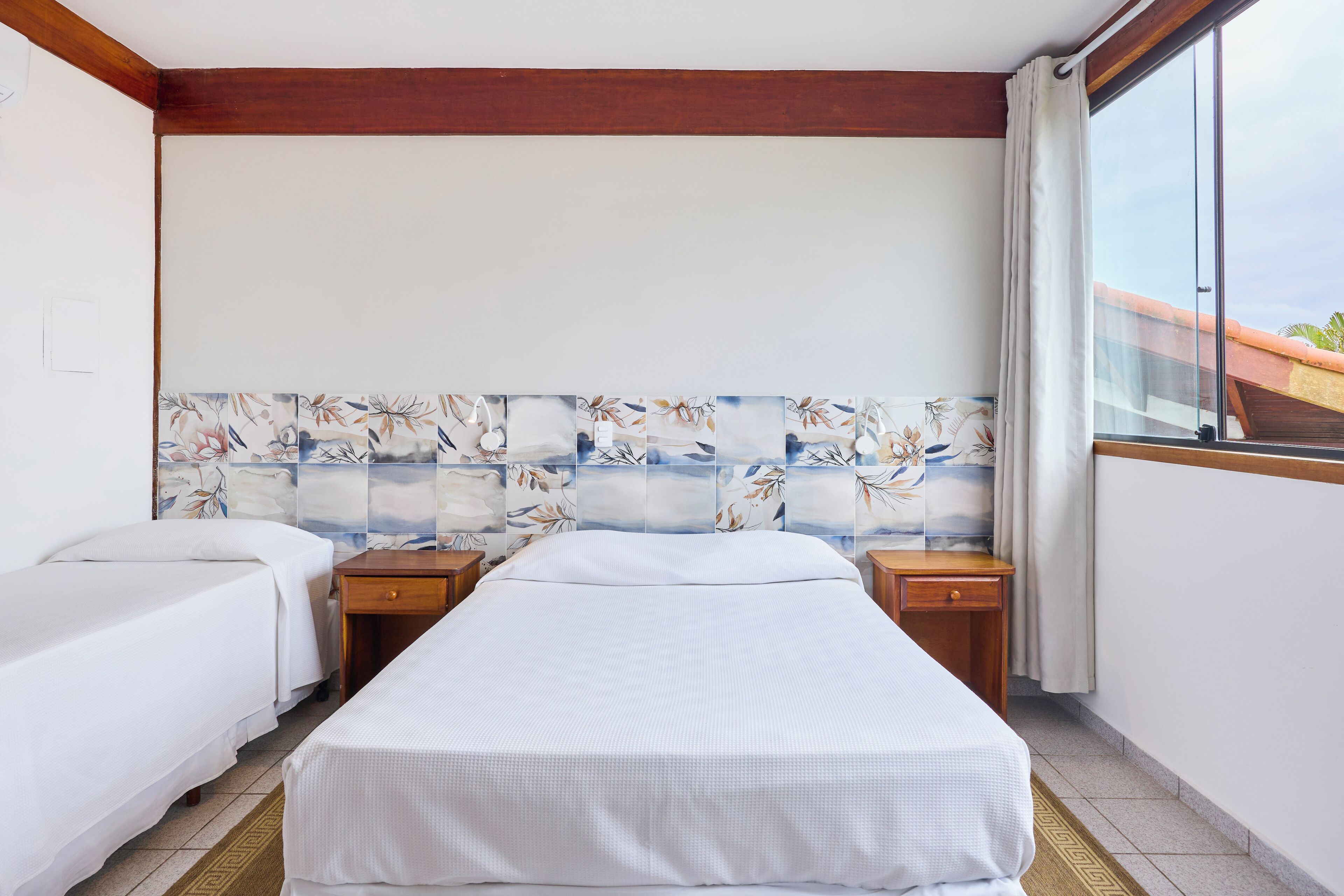 A hotel room with a large bed, two bedside tables, a window with curtains, and a decorative wall panel.